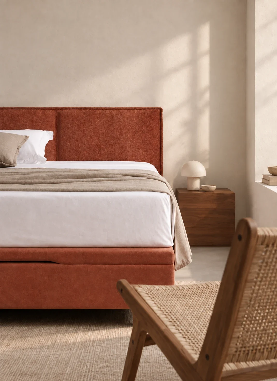Polsterbett mit terrakottafarbenem Kopfteil und gepolstertem Sockel in einem hellen Schlafzimmer, daneben ein Nachttisch aus Nussbaum und ein Geflechtstuhl